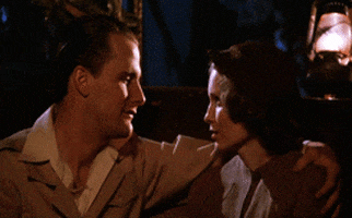 Movie gif. Jeff Daniels as Tom Baxter sits next to Mia Farrow as Cecilia in "The Purple Rose of Cairo," with his arm around her shoulder and leans in to kiss her.