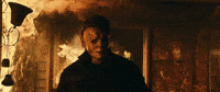 Movie gif. James Jude Courtney as Michael Myers in "Halloween Kills," walks toward us, away from a fire that rages out of a doorway behind him.
