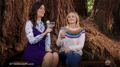 TV gif. D'Arcy Carden as Janet and Kristen Bell as Eleanor in The Good Place. The ladies are sitting on a tree stump and they clink their margaritas proudly.