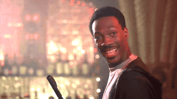 Movie gif. Eddie Murphy as Axel in Beverly Hills Cop looks over his shoulder and smiles with an open mouth as he holds up an enthusiastic OK with his hand. 