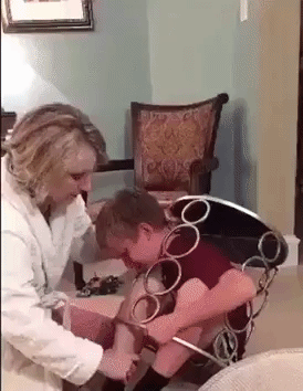 Video gif. Mother tries to free a scared boy stuck inside a metal-framed end table as his sister laughs hysterically.