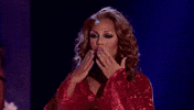 Reality TV gif. Glammed up contestant on RuPaul's Drag Race blows a kiss to the crowd and begins to strut away in her shimmering red long sleeve bikini top.