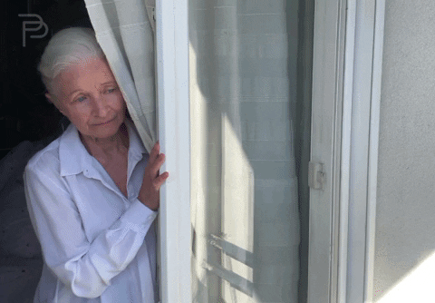 Video gif. An elderly woman looks out a window from behind a curtain, seeming lonely and sad. Sparkling pink text moves down the screen, "I miss you."