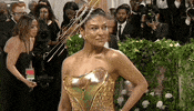 Met Gala 2024 gif. Closeup of Sabrina Harrison wearing a smooth gold-colored fitted bodice with a scooped sweetheart necklace that is filled with a melting Dali-esque clock down the front. She's wearing a gold bedazzled fascinator in the style of a clock face with rods extending from its center with numbers at the endpoints. 