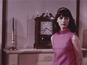 TV gif. A retro scene with washed-out colors, where a young woman in a sleeveless pink top looks impatiently at an antique clock.