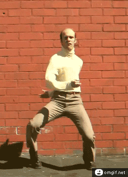 Video gif. Man wearing khakis and a cream-colored turtleneck dances in front of a brick wall, with his arms flailing like noodles.