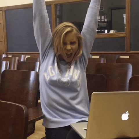 Video gif. A young woman sits in a classroom as she raises her hands in an excited cheer. 