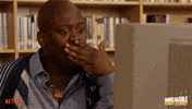 TV gif. Tituss Burgess as Titus Andromedon from Unbreakable Kimmy Schmidt covers his mouth in a shocked reaction to something on the screen of a library computer.