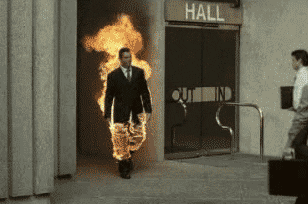 Video gif. Man in a suit and on fire walks nonchalantly as another man passing him shakes his hand, smiling warmly and not caring about the flames erupting from his body.