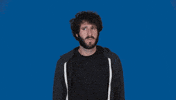 Celebrity gif. Lil Dicky stands in front of a solid blue background, looking from side to side with a slightly confused frown. He puts a hand to his chest as he speaks. Text, "Who me? He grins."