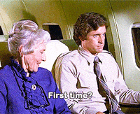 Movie gif. Robert Hays as Ted in Airplane! sits next to an old woman looking sick or nervous, hands locked to the rests and looking straight ahead. The old woman pats his hand reassuringly, smiling and asking, "First time?"