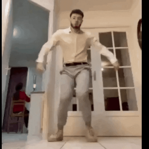 Video gif. A tall man wearing a white button down shirt and khaki pants awkwardly dances some variation of the Carlton dance while a woman sits in the background without acknowledging him. 
