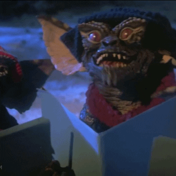 Movie gif. Dressed in scarves, earmuffs, and hats, a chorus of Gremlins hold up notebooks outside in the snow.