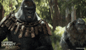 Trailer gif. A forest-set scene from the movie "Kingdom of the Planet of the Apes" shows a large, imposing metal-armored gorilla on horseback thrusting his right arm up and roaring in a war cry. A second chimpanzee on horseback can be seen behind him out of focus.