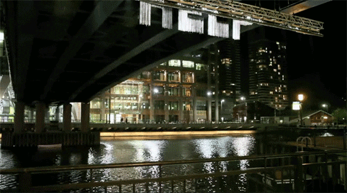 Video gif. Underneath a city bridge, water falls from a scaffold in the form of the word "Help," which then dissipates as it approaches the water.