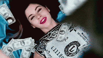 Video gif. A woman lays on the floor wearing a dollar bill dress. Cash money is poured over top of her like it’s raining dollar bills. She smiles happily and holds her hands out. 