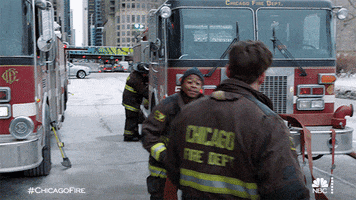 TV gif. Two firefighters from Chicago Fire walk by one another at the station and give each other a high five.