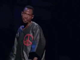 Celebrity gif. Martin Lawrence wearing a jacket with a peace sign on it, staring straight out and pointing at us with finger guns.