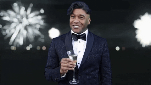 Celebrity gif. Wearing a tuxedo, Robert E Blackmon smiles and raises a flute of champagne to us as fireworks explode behind him.