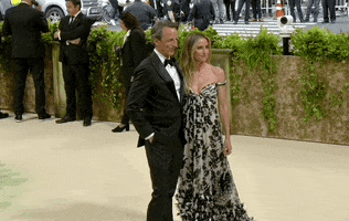 Met Gala 2024 gif. Seth Meyers poses with his wife Alexi Ashe on the red carpet, one hand in his pocket as he gives a big friendly smile at camera. Ashe is wearing a low-cut neutral beige dress with a black floral pattern and off-the-shoulder straps.