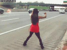 Video gif. A woman with long black hair dances a happy dance on the side of a busy interstate.
