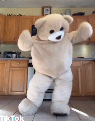 Video gif. Gigantic tan-colored teddy bear does a Fortnite dance in a kitchen, rocking his knees and flailing his arms.