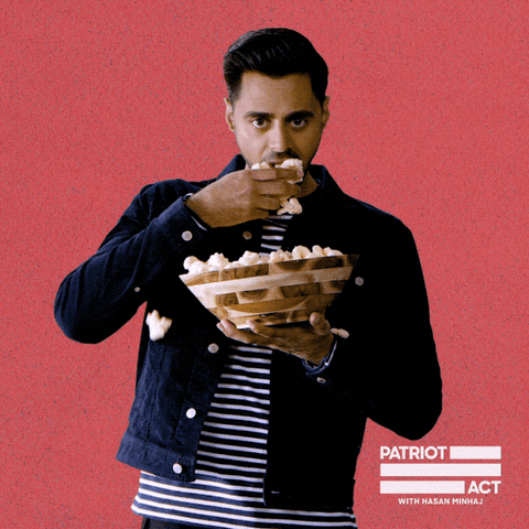 TV gif. Hasan Minhaj in Patriot Act locks eyes with us while holding a wooden bowl full of popcorn, shoving popcorn into his mouth as popcorn falls out into the bowl.