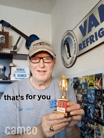 Celebrity gif. Robert R Shafer stands in a corner full of The Office memorabilia as he holds a trophy and taps it with his finger, smiling at us and saying, "That's for you."
