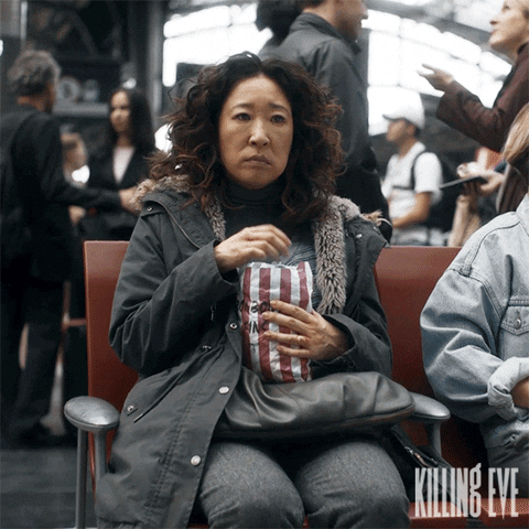 TV gif. Sandra Oh as Eve Polastri on Killing Eve sits in a busy train station, clutching a bag of popcorn. She nervously munches on the popcorn.