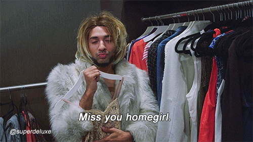 Video gif. Brandon Miller as Joanne the Scammer holds a gown as she stands between racks of clothing and tosses her head back while saying, "Miss you, homegirl." 