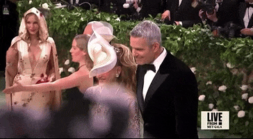 Met Gala 2024 gif. Sarah Jessica Parker poses with Andy Cohen. Parker is wearing a long sleeve scoop neck beige Richard Quinn dress with white and rhinestone detailing paired with a gold and white Philip Treacy fascinator that sweeps in a wave-like pattern diagonally across her face. Cohen is wearing a classic black tuxedo.
