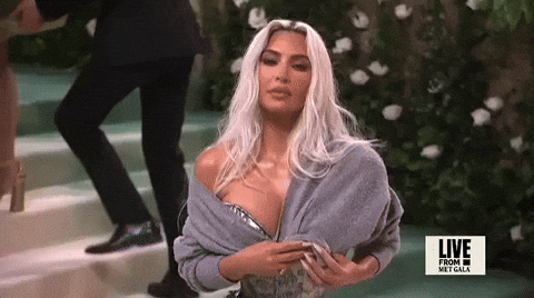 Met Gala 2024 gif. Met Gala 2024 gif. Kim Kardashian wearing a silver Maison Margiela gown and gray cardigan bolero, poses holding her sweater in place, twisting the end into her fingers.
