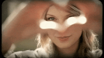Celebrity gif. Taylor Swift peeks through a heart shape made by her fingers before pointing at us with a smile. 
