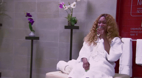 Reality TV gif. Tami Roman in Basketball Wives sits in her robe sipping on a flute of champagne. Something has tickled her pink and she is cackling in her seat, bending over and covering her mouth in laughter.