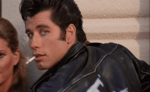 Movie gif. John Travolta as Danny Zuko in Grease looks over his shoulder and smiles with a cigarette in his mouth.