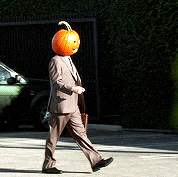 Video gif. A man wearing a suit and carrying a briefcase walking through a parking lot with a Jack-O-Lantern on his head.