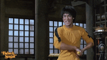 Movie gif. Wearing a yellow jumpsuit, Bruce Lee stands ready to fight, expertly throws nunchucks. From the movie Game of Death.