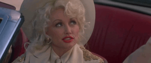 Movie gif. From The Best Little Whorehouse in Texas, Dolly Parton as Mona sits in the driver's seat of a convertible wearing a cowboy hat as she looks up and winks at someone with a growing smile. 