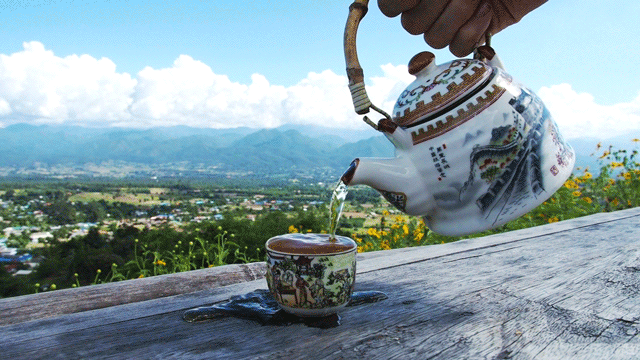 Digital art gif. Hand pours tea into a teacup, overflowing onto the wooden table overlooking scenery of a town and mountains.