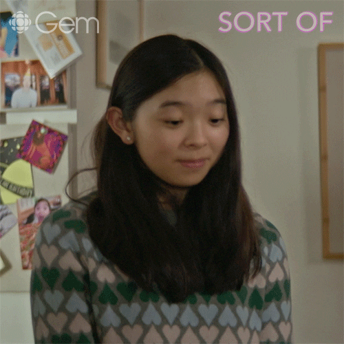 TV gif. Kaya Kanashiro as Violet in Sort Of. She looks down and shrugs her shoulders cheekily, acting as if she doesn't know what the person is referring to. 