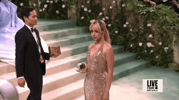 Met Gala 2024 gif. Camila Cabello turns to show the front of her Ludovic de Saint Sernin shimmery gold halter gown with a deep v-neckline. The dress is fitted through the waist with the skirt falling in a straight silhouette. The skirt has a high slit that comes to her upper thigh and she's holding a purse that is an actual rectangular block of ice with a rose inside.