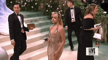 Met Gala 2024 gif. Camila Cabello turns to show the back of her Ludovic de Saint Sernin shimmery gold halter gown with a deep v-neckline. The dress is fitted through the waist with the skirt falling in a straight silhouette. Her hair is blonde with brown roots and styled in a long wavy ponytail that falls to her buttocks.