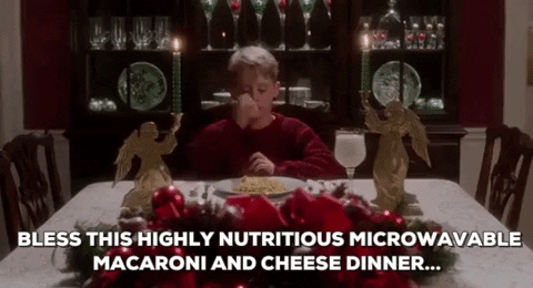 Movie gif. Macaulay Culkin as Kevin in Home Alone. He sits at the head of the table that's filled with Christmas decor and does the sign of the cross on himself as he says, "Bless this highly nutritional microwavable macaroni and cheese."