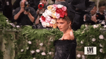 Met Gala 2024 gif. Zendaya wearing a big floral hat and throwing poses, lifting and dropping her chin with sultry eyes.