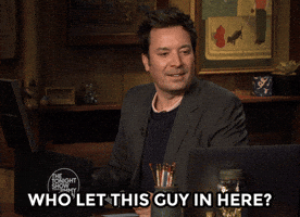 The Tonight Show gif. Jimmy Fallon looks down as if thinking hard as he says, “Who let this guy in here?” He then looks around, looking for an answer. 