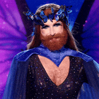 Reality TV gif. Drag Queen Gingzilla on the show Queen of the Universe with long lashes and a long beard wears a crown made up of blue butterflies, a blue cape, and a sparkly dress. They place their well manicured hand over their chest in appreciation and smile, nodding and batting away tears from their eyes.