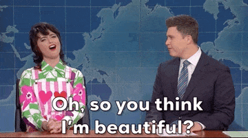 SNL gif. Melissa Villasenor and Colin Jost on the Weekend Update sit next to each other. Melissa looks up, batting her eyes, and holding her hands together firmly on the desk. She says, “Oh, so you think I'm beautiful?” Colin Jost laughs. 