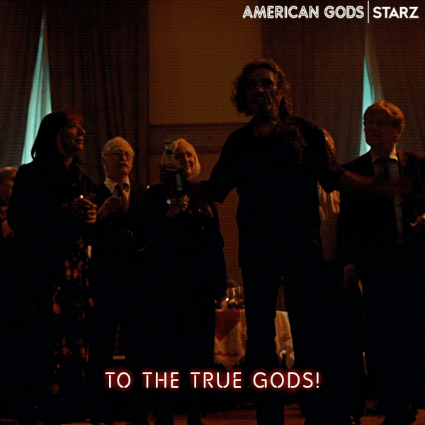 TV gif. Ian McShane as Mr. Wednesday on American Gods holds a bottle of alcohol up and yells, “To The True Gods!” Everyone lifts their glasses up and cheers with him.