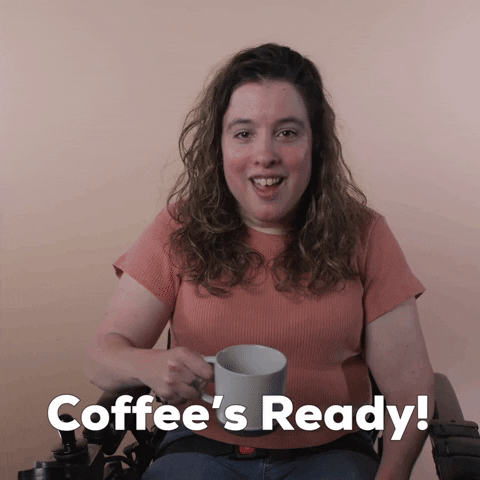 Reaction gif. A Disabled white-presenting Latina woman with cerebral palsy and kinky-curly brown hair, seated in her motorized wheelchair, gestures a mug in the air and says, "Coffee's ready!"
