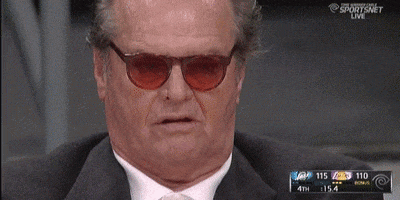 Celebrity gif. Jack Nicholson sits in the audience of a sports game. He wears tinted sunglasses and looks at the game with darting eyes. His mouth is slightly open as if bored.